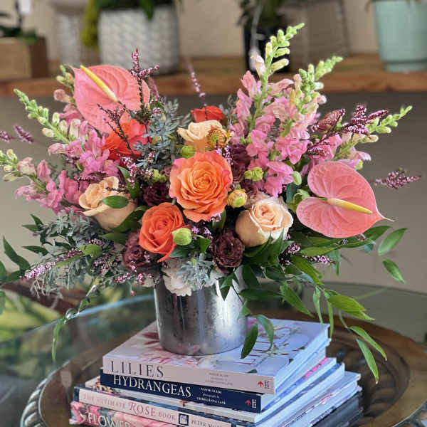 Colorful arrangement of peach roses, pink anthuriums, and snapdragons in a silver vase on books