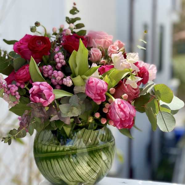 Bright pink and red tulips, roses, and ranunculus arranged in a round green glass vase