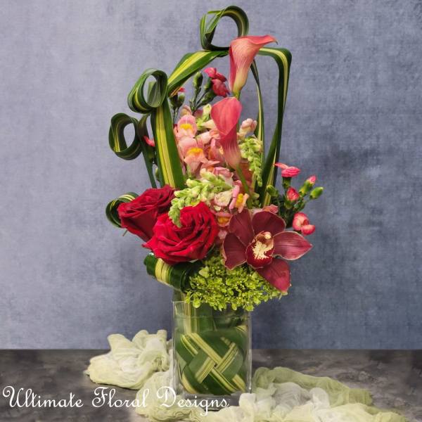 Tall floral arrangement with red roses, pink orchids, and curled green leaves in a glass vase
