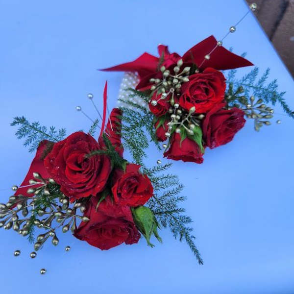 Two red rose wrist corsages with gold bead accents and ribbon on a light blue surface