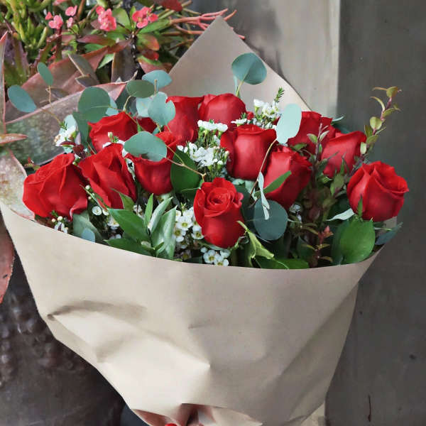 Hand-tied bouquet of red roses with small white flowers wrapped in kraft paper and tied with a red bow