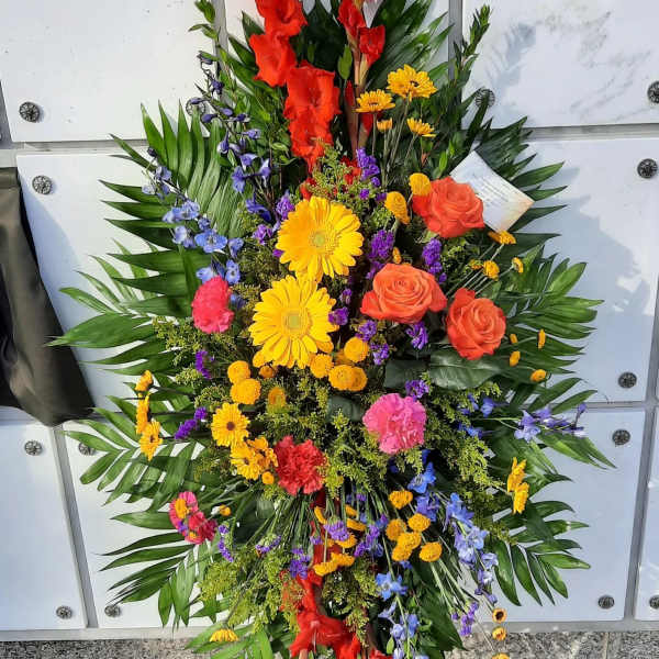 Tall standing spray of multicolored flowers including yellow gerbera, orange roses, and red gladiolus on an easel.