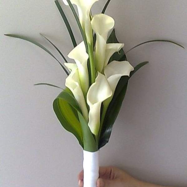 Hand-tied bouquet of white calla lilies with long linear foliage, wrapped in white ribbon.