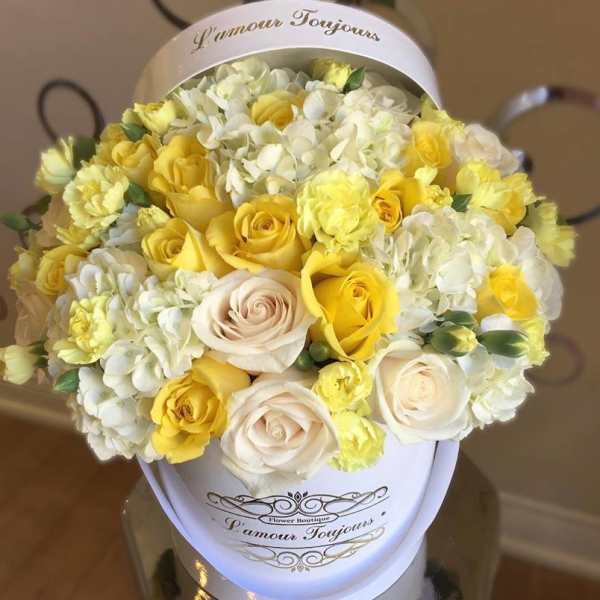 Round hatbox arrangement of yellow and cream roses, carnations, and white hydrangeas in a white container