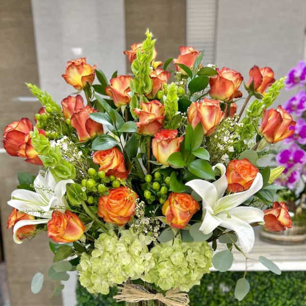 Tall arrangement of orange roses, white lilies, and green hydrangeas in a clear glass vase
