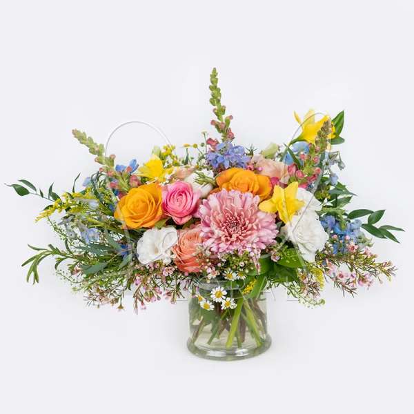 Low glass vase of mixed pink, yellow, white, and blue flowers in a lush garden-style arrangement