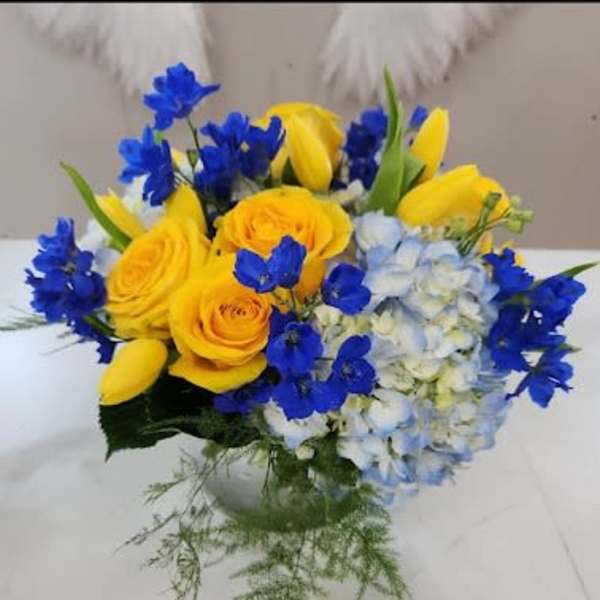 Low arrangement of yellow roses and tulips with blue delphinium and hydrangea in a clear glass vase
