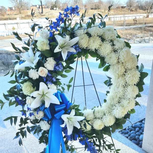 Standing white and blue funeral wreath with lilies and ribbon on a metal easel.