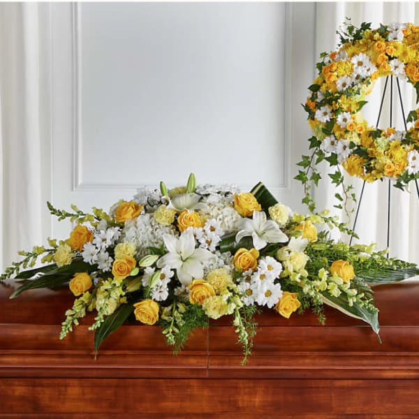 Yellow and white casket spray with roses, lilies, and daisies on a wood casket with a matching wreath behind it
