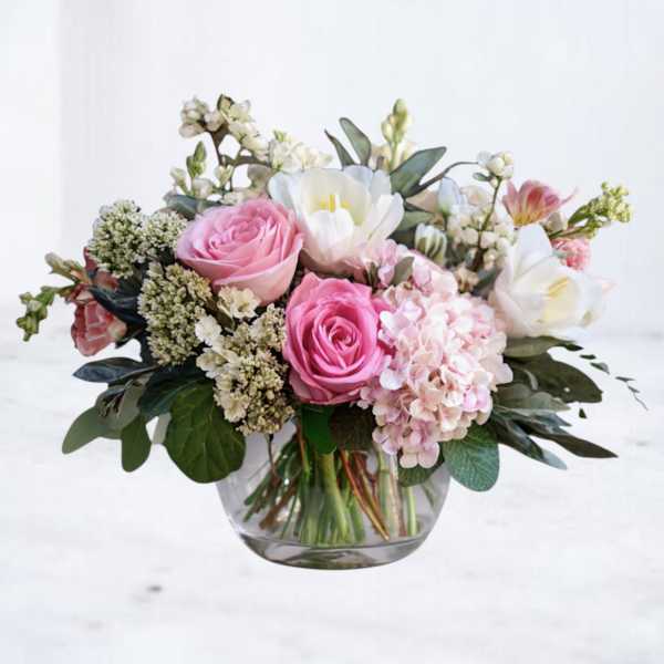 Low vase arrangement of pink roses, white blooms, and blush hydrangea in a clear glass bowl.