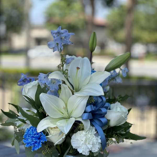White lilies and blue flowers in a clear glass vase with a blue ribbon