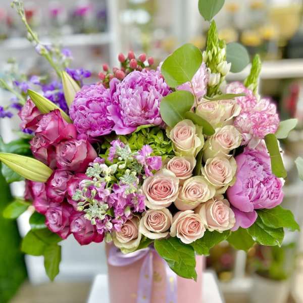 Pink and blush roses, peonies, and lilies arranged in a round pink hatbox with ribbon