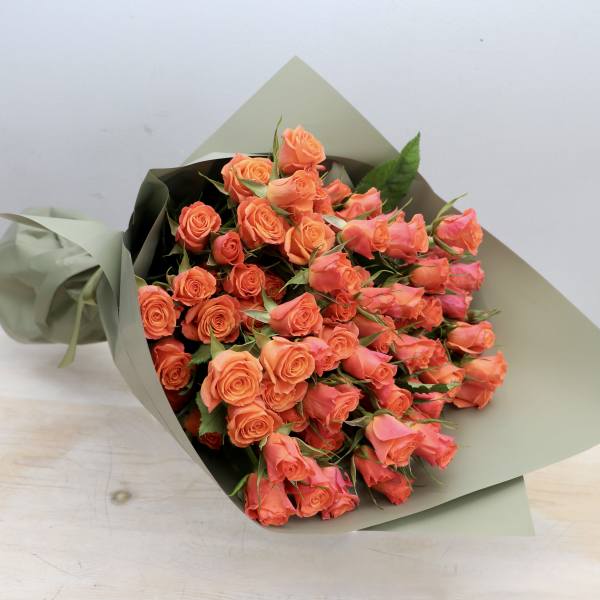 Bouquet of bright orange roses wrapped in matte sage green paper, resting on a light wooden surface