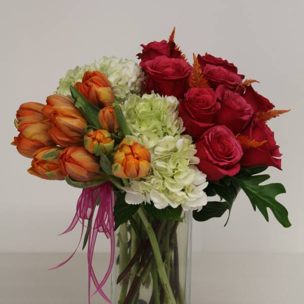 Tall glass vase with red roses, orange tulips, and pale green hydrangeas accented with stones at the base