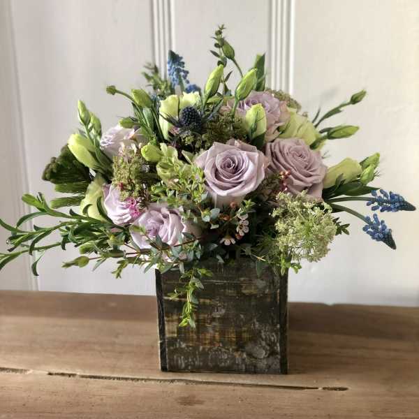 Lavender roses and white buds arranged with blue accents in a rustic wooden cube vase