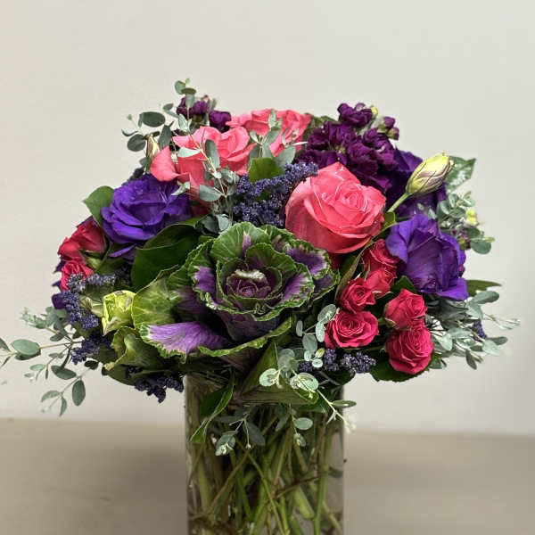 Vibrant arrangement of pink roses, purple flowers, and ornamental cabbage in a clear glass vase