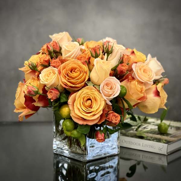 Low arrangement of orange and peach roses in a glass cube vase on a reflective table