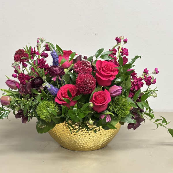 Low arrangement of hot pink roses and mixed purple flowers in a hammered gold bowl.