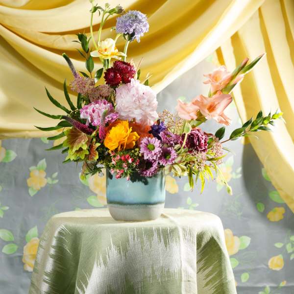 Colorful mixed flower arrangement in a blue ceramic vase on a draped table with yellow fabric backdrop.