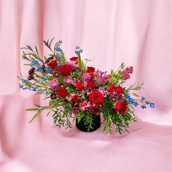 Asymmetrical arrangement of red and pink roses with blue accents in a black vase on a pink backdrop
