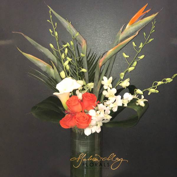 Tropical bouquet with orange roses, white calla lilies, and orchids in a tall glass vase