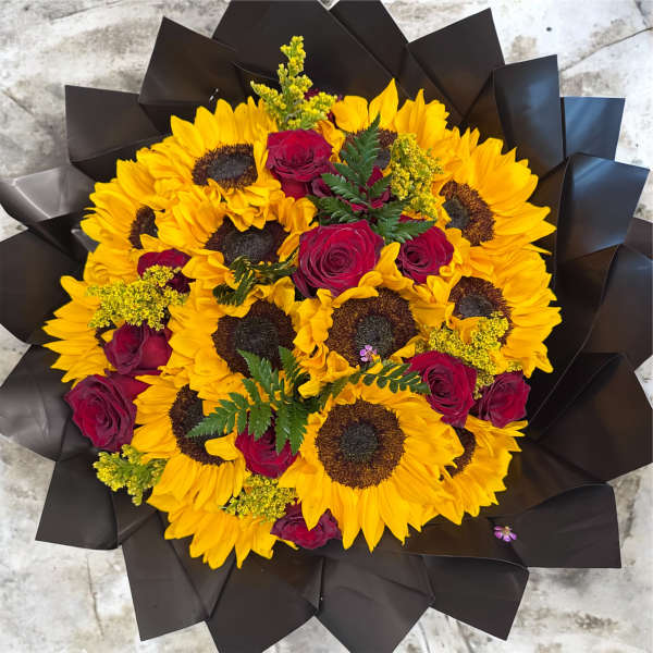 Round bouquet of bright yellow sunflowers and red roses wrapped in black paper.