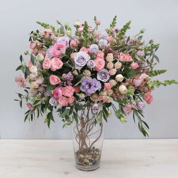 Tall arrangement of pink and lavender roses and lisianthus in a clear glass vase with stones