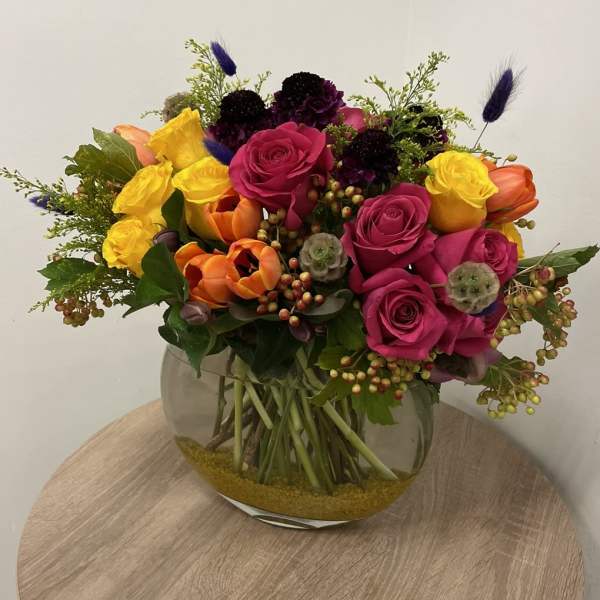 Bright arrangement of pink and yellow roses, orange tulips and purple flowers in a round glass vase.