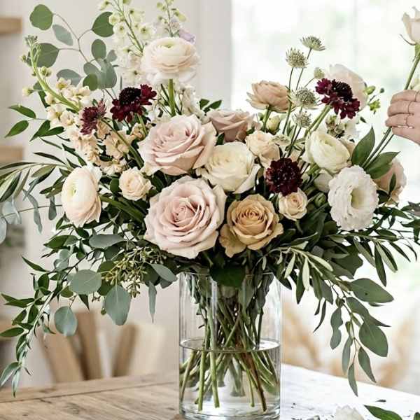 Large blush and cream rose arrangement with burgundy accents in a clear glass vase on a table