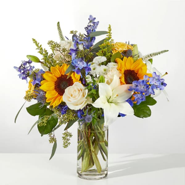Bouquet of sunflowers, white lilies, and blue flowers in a clear glass vase