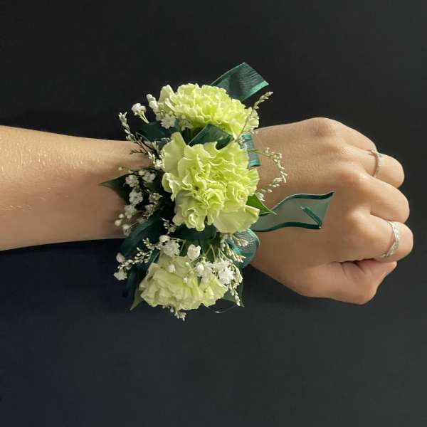 Light green carnation wrist corsage with white filler flowers and dark green ribbon on a hand.