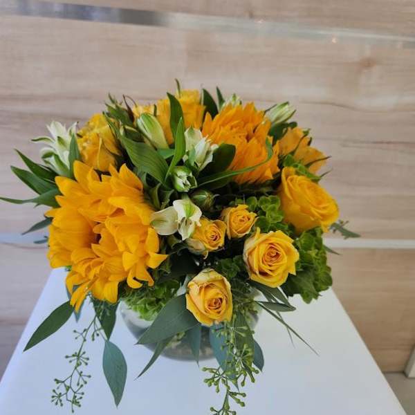 Low arrangement of yellow roses and yellow flowers in a round glass vase on a white table