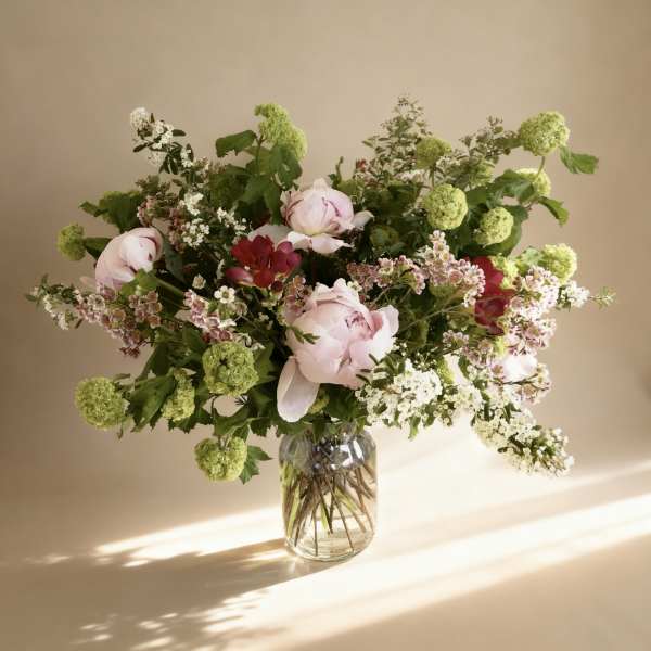 Loose arrangement of pale pink peonies, red blooms, and green flower clusters in a clear glass vase.