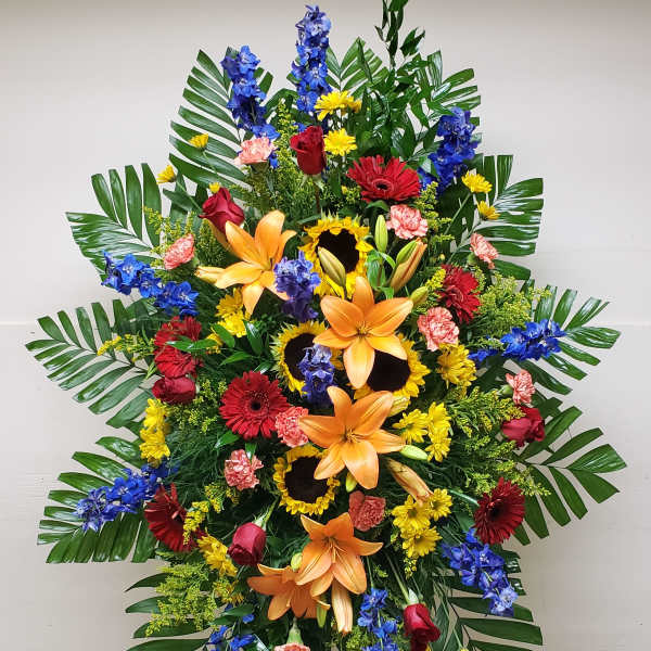 Tall standing spray of orange lilies, sunflowers, red roses, and mixed bright flowers on an easel.