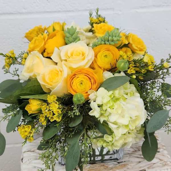 Low yellow and cream flower arrangement with roses, ranunculus, hydrangea, and succulents in a glass cube vase