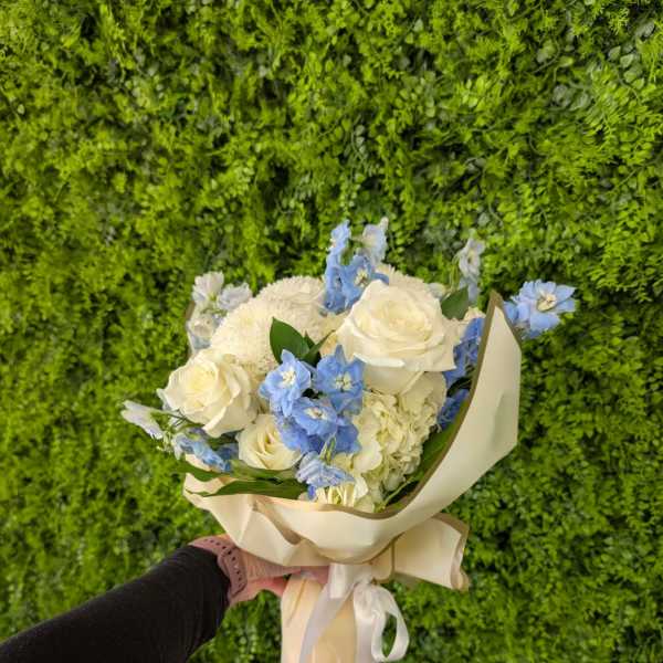 Hand-tied bouquet of white roses and blue flowers in cream wrap with ribbon, held against a green wall.