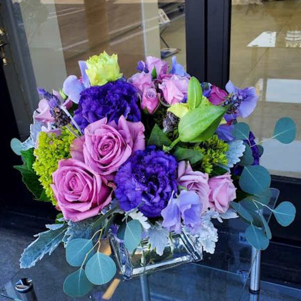 Low arrangement of pink roses and purple flowers in a square glass vase on a glass table.