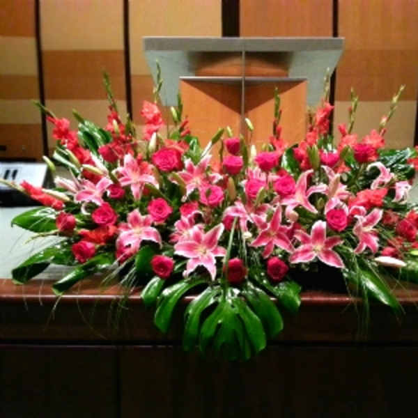 Large altar spray of pink lilies and red roses with bold tropical leaves on a wooden podium.