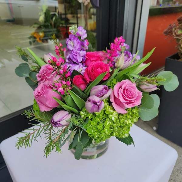 Low pink and purple flower arrangement with green hydrangea in a clear glass vase on a white table