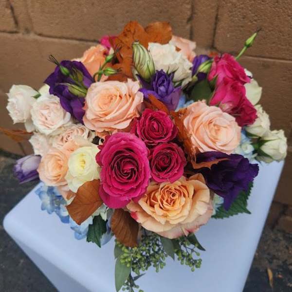 Round bouquet of pink and peach roses with purple lisianthus and blue accents on a white pedestal