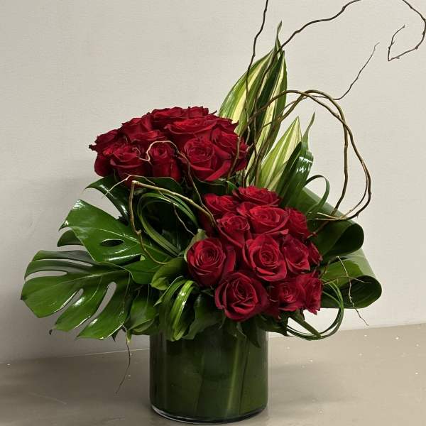 Tall arrangement of red roses with tropical green leaves and curly branches in a clear glass cylinder vase