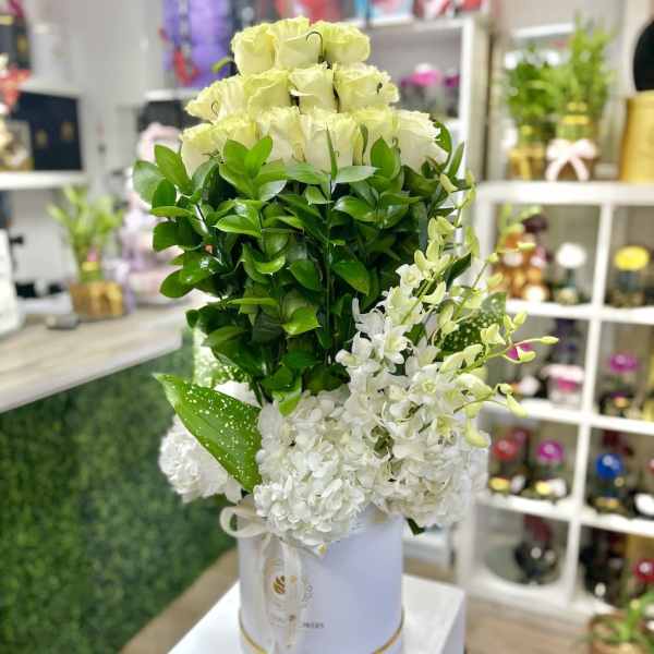 Tall arrangement of pale roses, white blooms, and dense greenery in a white hatbox container