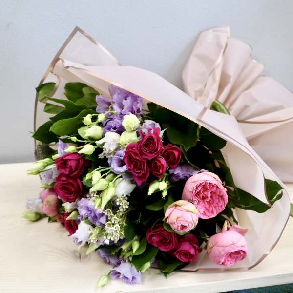 Pink and lavender bouquet of roses and lisianthus wrapped in blush paper, resting on a light wood surface