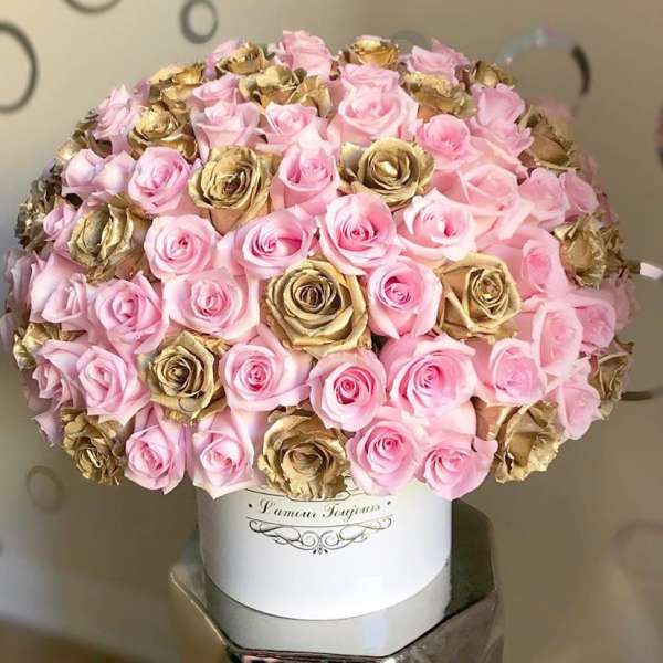 Dome of pink and gold roses arranged densely in a white hatbox-style container on a pedestal