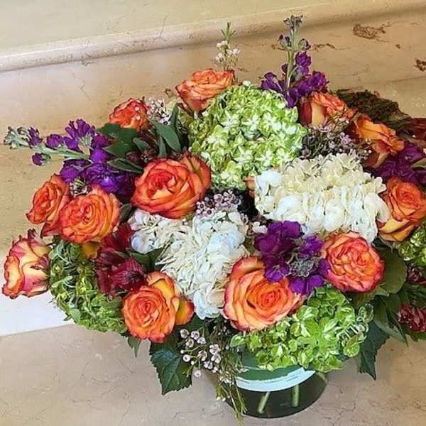 Low round arrangement of orange roses, green and white hydrangeas, and purple blooms in a glass vase