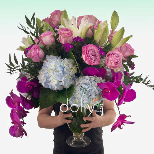 Tall arrangement of pink roses, white lilies, blue hydrangeas, and fuchsia orchids in a clear glass vase