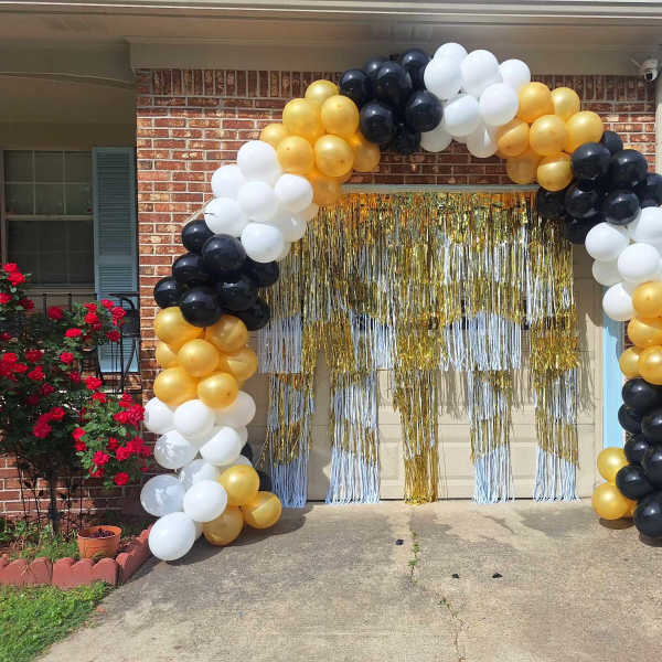 Black, white, and gold balloon arch framing a garage doorway with gold metallic fringe curtains.