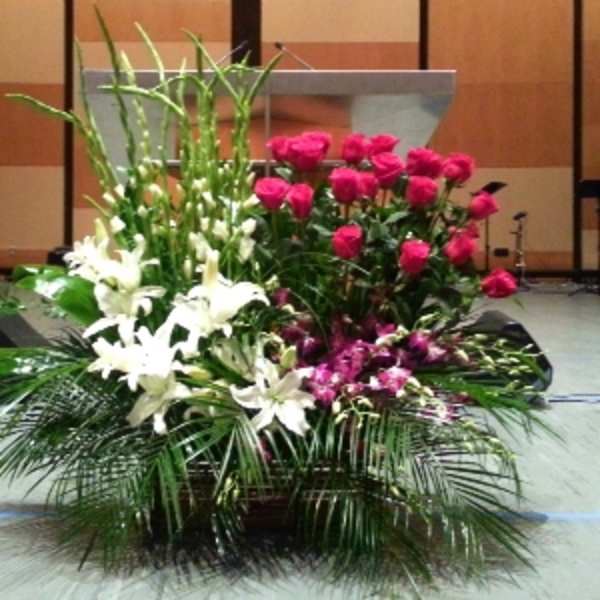 Large stage arrangement of white lilies, hot pink roses, and purple flowers with tall green spikes in a low container.
