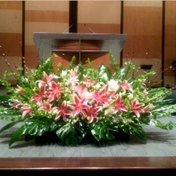 Large low arrangement of pink lilies and lush foliage in front of a podium on a stage