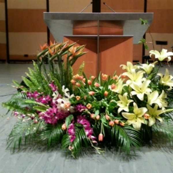 Large mixed floral arrangement with yellow lilies, orange tulips, and purple flowers in front of a podium